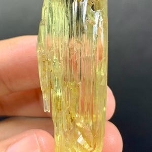 May include: A translucent, yellow-green crystal with a rough, textured surface. The crystal is held in a hand, showcasing its elongated, columnar structure and natural facets. The colour is a vibrant, pale yellow.