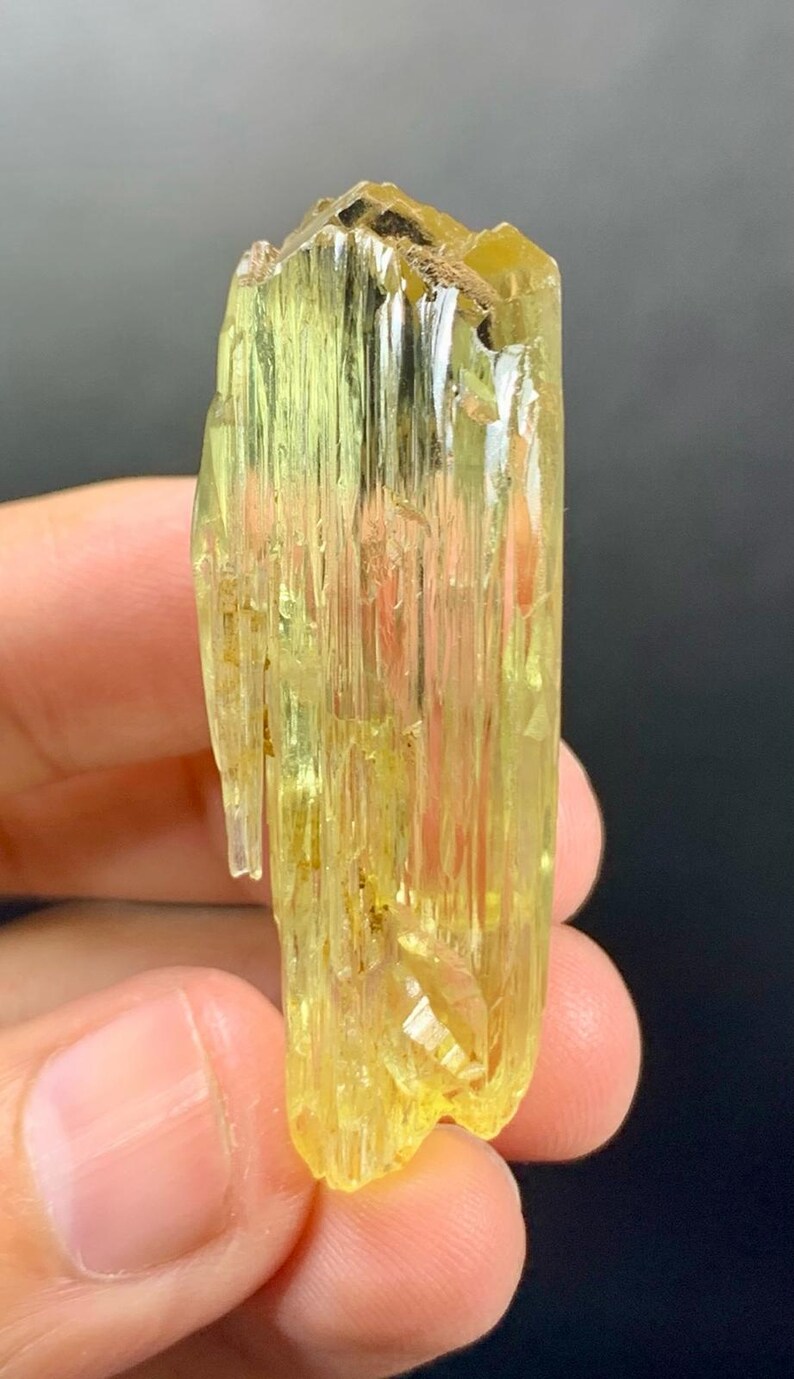 May include: A translucent, yellow-green crystal with a rough, textured surface. The crystal is held in a hand, showcasing its elongated, columnar structure and natural facets. The colour is a vibrant, pale yellow.