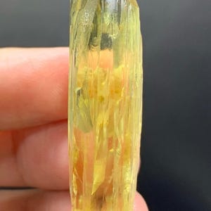 May include: A yellow crystal with a rough, textured surface. The crystal is held between fingers, showcasing its elongated, columnar shape and natural, unpolished appearance. The colour ranges from pale to vibrant yellow.