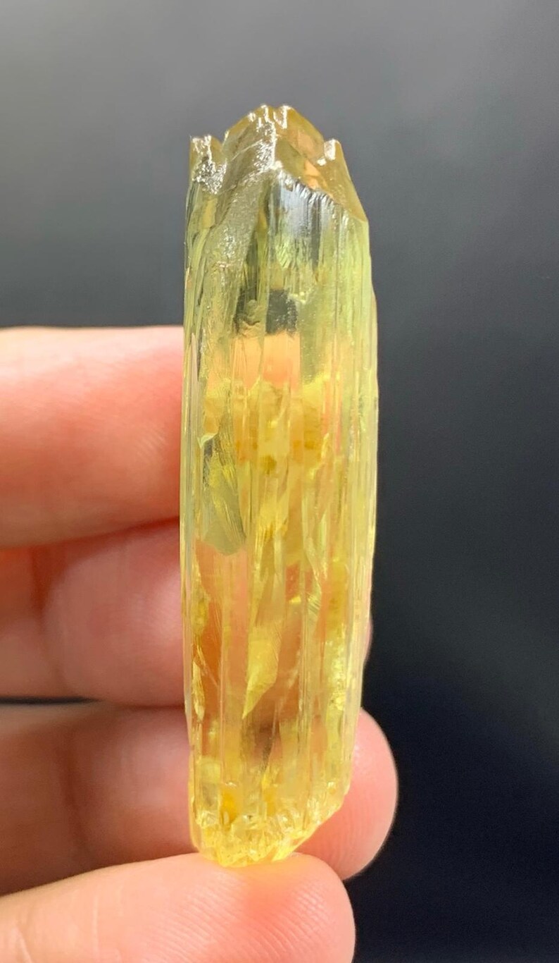 May include: A yellow crystal with a rough, textured surface. The crystal is held between fingers, showcasing its elongated, columnar shape and natural, unpolished appearance. The colour ranges from pale to vibrant yellow.