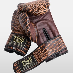 May include: A pair of brown leather boxing gloves with a crocodile pattern. The gloves have black accents and a gold logo that reads "TSSB SPORTS" with the website address "www.tssbsports.com".