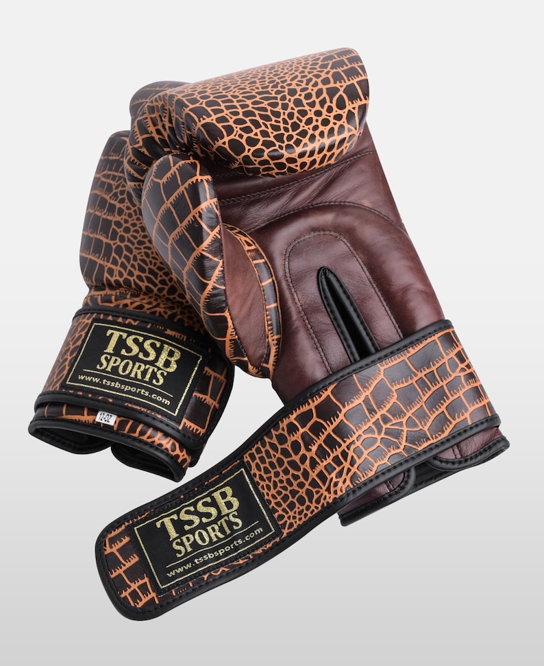 May include: A pair of brown leather boxing gloves with a crocodile pattern. The gloves have black accents and a gold logo that reads "TSSB SPORTS" with the website address "www.tssbsports.com".