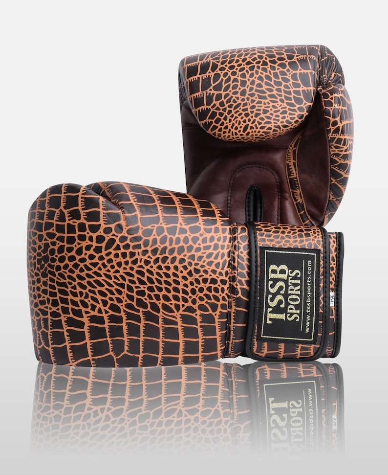 May include: A pair of brown and black boxing gloves with a crocodile skin pattern. The gloves have a black strap with the text "TSSB SPORTS" and "www.tssbsports.com" printed on it.