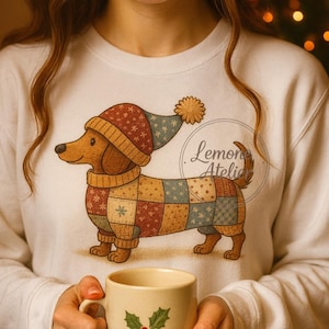Puede incluir: Sudadera blanca con una ilustración de un perro salchicha con un suéter de retazos y un gorro de invierno. El perro está representado en marrón, rojo, azul y amarillo. La sudadera tiene un cuello redondo. La ilustración tiene el texto "Lemon Atelier".