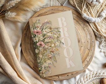 Könnte beinhalten: Ein beiger Notizblock mit einer Blumenillustration und dem Wort "BROOK" in rosa Schrift.