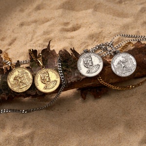 Puede incluir: Cuatro colgantes de oro y plata con cadenas, que presentan diseños de inspiración romana. Los colgantes están sobre una superficie de madera con un fondo arenoso.