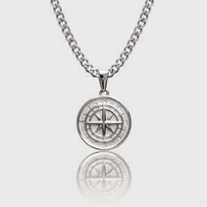 May include: Silver compass pendant necklace. The round pendant features a detailed compass rose design with directional markers. The necklace has a thick, textured chain. The pendant is reflected in the surface below.