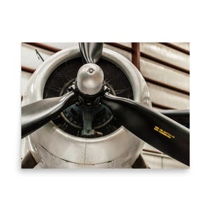 May include: A silver aeroplane engine cowling with a central propeller hub and three black propeller blades. The blades feature yellow text reading "DEG.NO.6477A-12" and "SER.NO.8880082". A dark metal mesh screen is visible behind the propeller assembly.