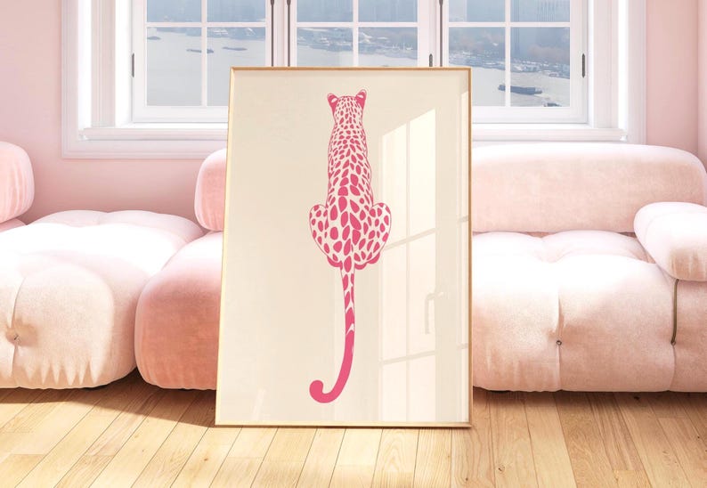 May include: A framed art print featuring a pink leopard illustration. The leopard is depicted from the back, with a long tail and spotted pattern, set against a cream background. The artwork is displayed in a room with pink furniture and a wooden floor.