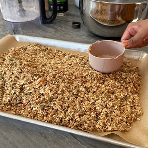 May include: A baking tray lined with baking parchment, filled with a mixture of granola, nuts, and seeds. The granola is a light brown colour and has a slightly crunchy texture. The nuts and seeds are a variety of colours and sizes. The baking tray is on a grey countertop.