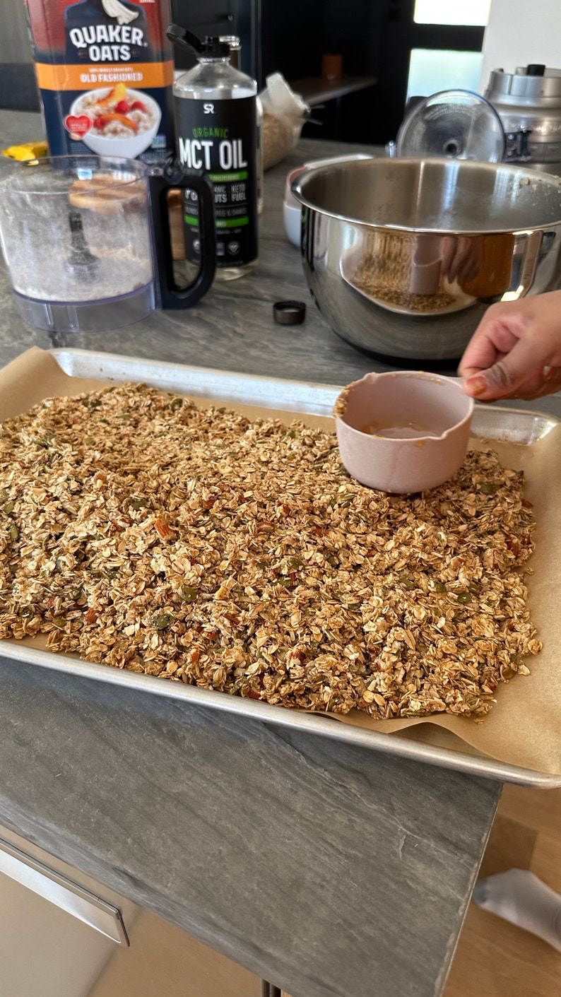 May include: A baking tray lined with baking parchment, filled with a mixture of granola, nuts, and seeds. The granola is a light brown colour and has a slightly crunchy texture. The nuts and seeds are a variety of colours and sizes. The baking tray is on a grey countertop.
