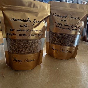 May include: Two gold bags filled with homemade granola. The bags are labelled "Homemade Granola with Oats, almond, coconut oil, pumpkin seed, maple syrup" and "Homemade Gran with oats, almond, coconut oil, pumpkin seeds, maple syrup". Both bags have the text "Merry Christmas" written on them.