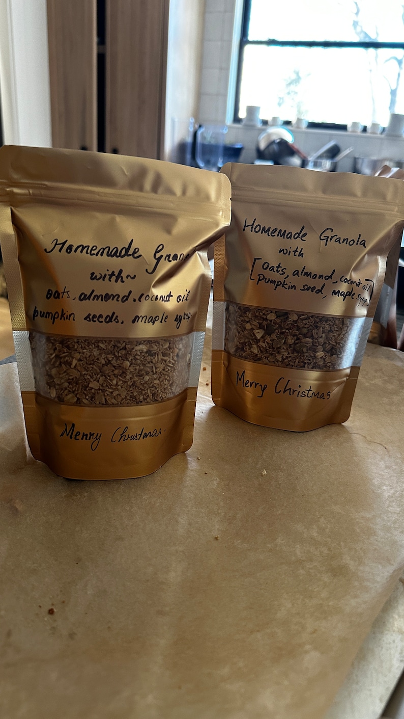 May include: Two gold bags filled with homemade granola. The bags are labelled "Homemade Granola with Oats, almond, coconut oil, pumpkin seed, maple syrup" and "Homemade Gran with oats, almond, coconut oil, pumpkin seeds, maple syrup". Both bags have the text "Merry Christmas" written on them.