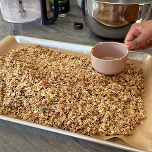 May include: A baking tray lined with baking parchment, filled with a large batch of granola. The granola is a mixture of oats, nuts, seeds, and dried fruit. A pink measuring cup is on the right side of the tray.