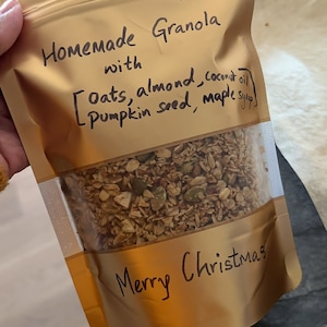 May include: A gold resealable bag filled with homemade granola. The bag has a handwritten label that reads "Homemade Granola with [Oats, almond, coconut oil, pumpkin seed, maple syrup] Merry Christmas".