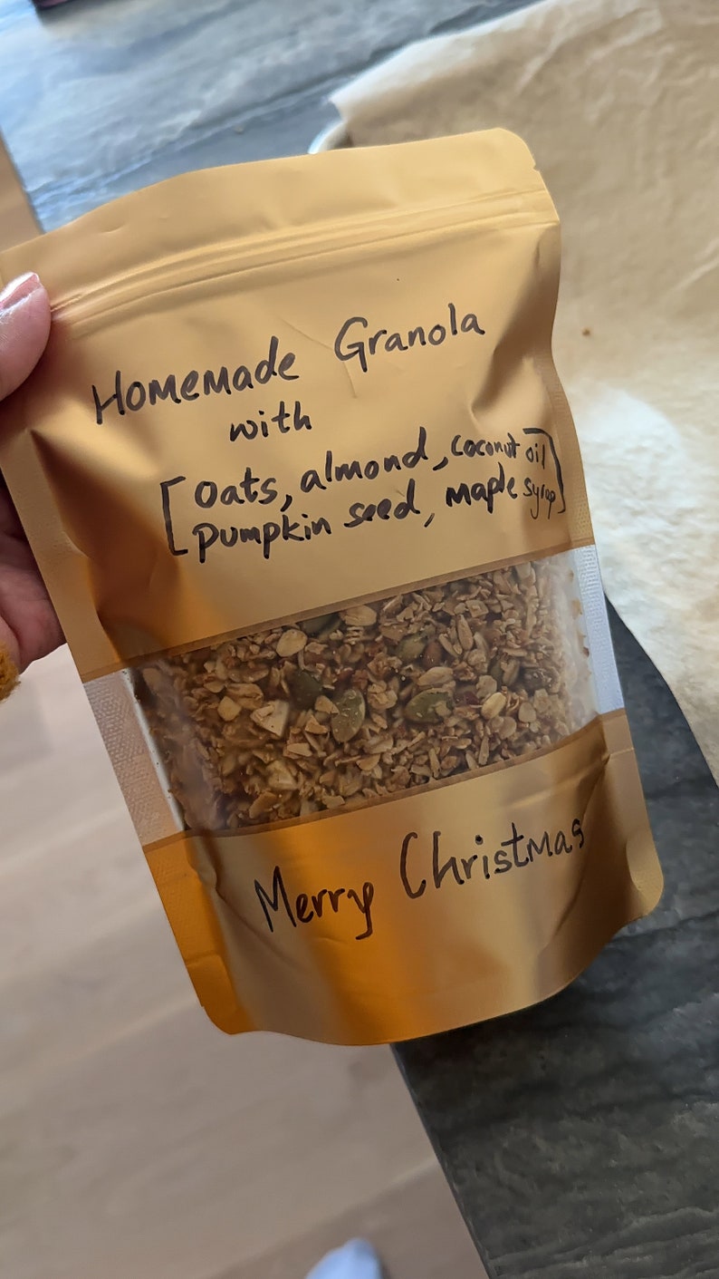 May include: A gold resealable bag filled with homemade granola. The bag has a handwritten label that reads "Homemade Granola with [Oats, almond, coconut oil, pumpkin seed, maple syrup] Merry Christmas".