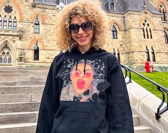 Puede incluir: Una sudadera con capucha negra con un estampado gráfico de una mujer con gafas de sol rosas y la lengua fuera. La mujer está rodeada de un diseño abstracto en blanco y negro.