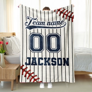 May include: White and blue baseball jersey fleece blanket with red stitching. The blanket has a number "00" and the name "JACKSON" printed on it. The text "Team name" is printed in a curved font at the top of the blanket.