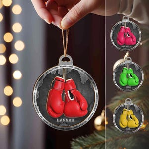 May include: A silver Christmas ornament with a boxing glove design. The ornament features a pair of red boxing gloves with the name "HANNAH" inscribed on the ornament. The ornament is surrounded by a circle of words that describe the qualities of a boxer, such as "RESILIENT", "BRAVE", "FOCUSED", "STRONG", "QUICK", "COMMITTED", "COURAGEOUS", "PERSISTENT", "PASSIONATE", "HONEST", "ATHLETE", "ATTITUDE", "MOTIVATED", "DREAMS", "BELIEVE", "FIGHT", "WIN", "NEVER GIVE UP".