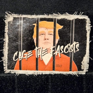 May include: A rectangular patch with a distressed edge, featuring an illustration of a person behind bars. The person has orange hair and is wearing an orange jumpsuit. The text "CAGE THE FASCISTS" is written across the top.
