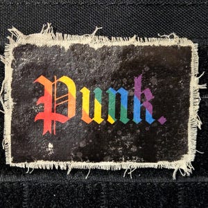 May include: A rectangular patch with the word "Punk" in rainbow-colored gothic lettering. The patch has a black background with white speckles and a frayed, off-white border. The patch is likely for clothing or accessories.