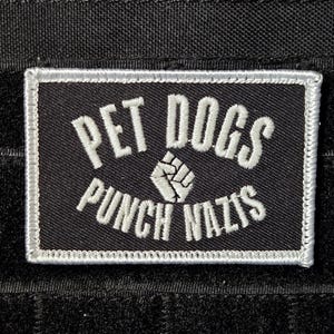 May include: Black rectangular patch with white embroidered text that reads "PET DOGS PUNCH NAZIS". The patch has a white border and a clenched fist graphic. The background is a textured black fabric.
