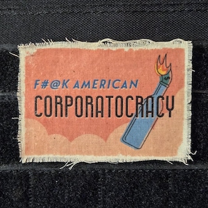 May include: A red, white, and blue patch with the text "F#@K AMERICAN CORPORATOCRACY" and a cartoon image of a lit match. The patch is made of fabric and has a frayed edge.