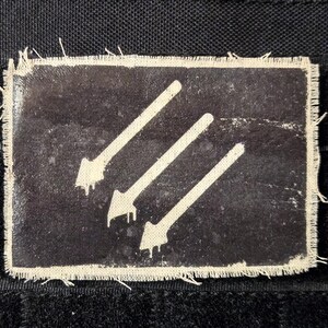 May include: A rectangular patch with a black background and three white arrows pointing diagonally. The arrows have a distressed, dripping effect at the tips. The patch has a frayed, off-white border and is set against a black fabric.