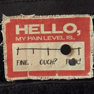 May include: Red fabric patch with white text that reads "HELLO, MY PAIN LEVEL IS..." with a black marker line and a black dot on the line. The line has the words "FINE", "OUCH?" and "F*K!" written below it.