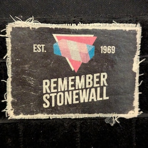 May include: A black and white patch with a pink and blue triangle design. The patch says "EST. 1969" and "REMEMBER STONEWALL".