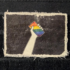 May include: A rectangular patch with a distressed black background and a frayed edge. The patch features a hand holding a rainbow-colored block. The design is a symbol of LGBTQ+ pride and support.