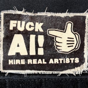 May include: A rectangular black fabric patch with frayed edges featuring white printed text that reads FUCK AI! HIRE REAL ARTISTS and a white graphic illustration of a hand making a finger gun gesture pointing toward the left side.