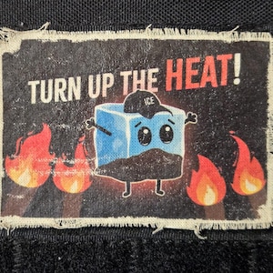 May include: A rectangular patch with a distressed border featuring the text "TURN UP THE HEAT!" above a cartoon ice cube wearing a hat that says "ICE". Flames are on either side of the ice cube.