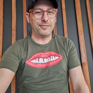May include: An olive green t-shirt featuring a red lip graphic with the word "SPEECH?" in white. The person in the image is wearing a black baseball cap and red-framed glasses.