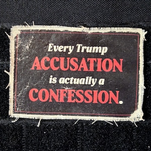 May include: A rectangular patch with frayed edges, featuring the text "Every Trump ACCUSATION is actually a CONFESSION" in white and red on a black background. The patch is set against a black textured surface.