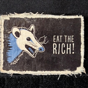 May include: A rectangular patch with a black background and frayed edges. It features a cartoon opossum with blue fur and the text "EAT THE RICH!" in white. The opossum is facing left with its mouth open.