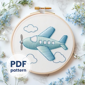 May include: A blue airplane with a propeller embroidered on a white fabric in a wooden embroidery hoop. The airplane is flying in the sky with clouds. The text "PDF pattern" is below the hoop.
