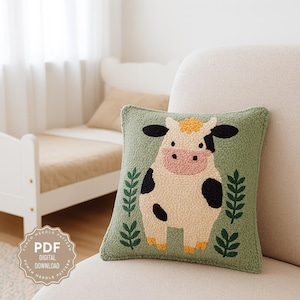 May include: A square, green decorative cushion featuring a punch needle design of a cow. The cow has black spots, ears, and a yellow and beige body. The cushion has a leaf design on each side. The text "PDF DIGITAL DOWNLOAD" is in a circle on the bottom left.