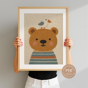 May include: A framed artwork featuring a friendly brown bear with a blue and orange striped shirt, and a small bird perched on its head. The artwork has a textured, beige background and is held by a person.