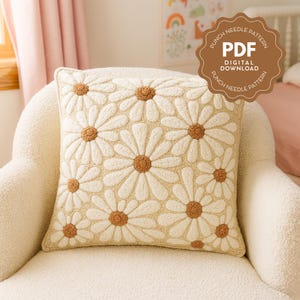 Op de afbeelding: Crèmekleurig vierkant sierkussen met een madeliefjespatroon. De madeliefjes hebben witte bloemblaadjes en bruine centra. Het kussen ligt op een crèmekleurige stoel. Een bruin label rechtsboven leest "PUNCH NEEDLE PATTERN PDF DIGITAL DOWNLOAD".