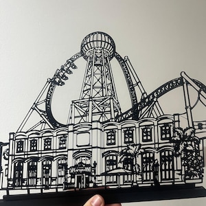 Könnte beinhalten: Eine schwarze, lasergeschnittene Metallsilhouette, die eine Achterbahnstruktur mit einem zentralen Turm und einer Gebäudefassade mit Rundbogenfenstern, Palmen und dem Schriftzug "VORTEX" über dem Haupteingang darstellt.