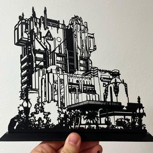 May include: A black, flat, laser-cut silhouette of a large, multi-story building with intricate architectural details, including pipes, towers, and arched structures, mounted on a rectangular base, depicting a complex industrial-style facade.