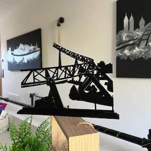May include: A black metal sculpture of a bridge, featuring intricate details of the structure. The sculpture is displayed on a wooden block, with a plant in a black pot in the foreground. Artwork hangs on the white wall in the background.