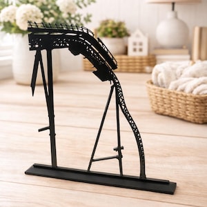 Könnte beinhalten: Schwarze Metall-Achterbahn-Skulptur. Das dekorative Stück zeigt ein detailliertes Design einer Achterbahnstrecke mit Stützen und einem Sockel. Die Skulptur ist ein einzigartiges Wohnaccessoire.