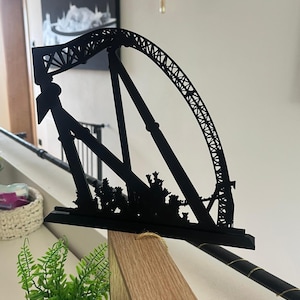 May include: A black metal sculpture of a roller coaster, featuring a curved track and support structure. The sculpture is placed on a wooden surface, with a decorative plant in the foreground. The roller coaster design includes intricate details.