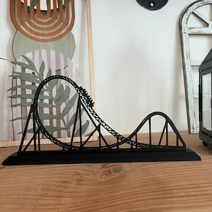 Może przedstawiać: Czarny miniaturowy model kolejki górskiej. Kolejka górska ma pętlę i wzniesienie, a tor jest podparty ukośnymi belkami. Model jest eksponowany na drewnianej powierzchni, prezentując swój skomplikowany design.