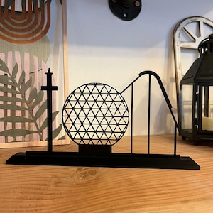 Könnte beinhalten: Eine schwarze Metallskulptur einer Achterbahn mit einer geometrischen Kugel und einer hohen, dünnen Struktur. Die Skulptur steht auf einer Holzoberfläche, mit einem dekorativen Stück und einer Laterne im Hintergrund.