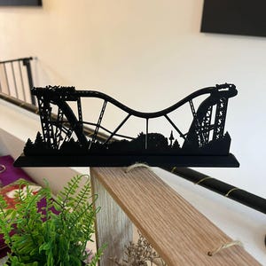May include: A black, three-dimensional model of a roller coaster. The model features intricate details of the track, supports, and surrounding landscape. The coaster design includes a series of hills and drops, showcasing a thrilling ride. The model is displayed on a wooden surface.