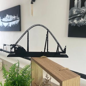 May include: A black metal roller coaster sculpture. The sculpture depicts the track and supports of a roller coaster, with a loop and various hills. The sculpture is displayed on a wooden shelf, with artwork in the background.
