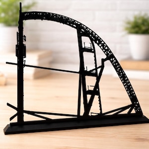 Può includere: Una scultura di ponte in metallo nero con un design ad arco. La scultura è una silhouette, che mostra la struttura del ponte, compreso l'arco, le travi di supporto e la base. Si trova su una superficie di legno.